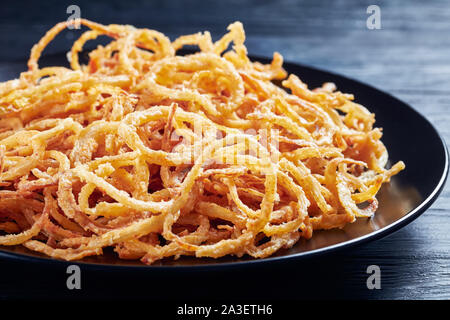 close-up of french crispy fried onion rings and strings on a black plate on a wooden table, horizontal view from above, macro Stock Photo