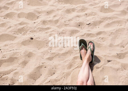 Female feet with legs crossed wearing a pair of flip flops unidentifed as we look down on a sandy beach. Stock Photo