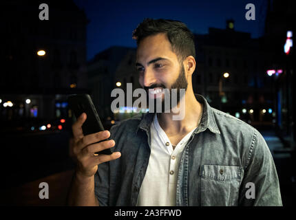 Young bearded smiling arab man holding smartphone outdoors night street Stock Photo