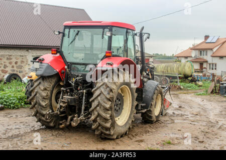 Red  tractor and agricultural equipment in the yard of a dairy farm. Back view. A barn and an apartment building in the background . Podlasie, Poland. Stock Photo