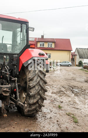 Red tractor  in the courtyard of a dairy farm. View from the back. Apartment building and car in the background. Podlasie, Poland. Stock Photo