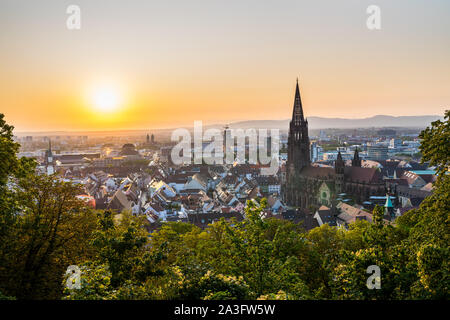 Germany, Romantic orange sunset sky decorating skyline of city freiburg im breisgau houses and cathedral seen from above, aerial view over tree tops a Stock Photo