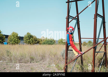 a scary clown wearing a colorful yellow, red and blue costume outdoors, hanging from the rusty structure of an abandoned billboard Stock Photo