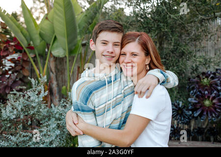 Smiling mom hugging teen son with lush plants on background Stock Photo