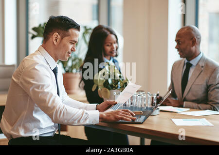 Smiling businessman reading graphs on a piece of paper and working on a laptop while sitting at an office table with diverse colleagues Stock Photo