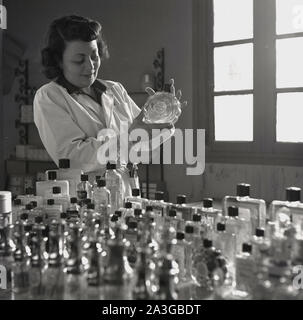 1950s, historical, Grasse, at a perfumery, a female chemist in a white-coat holding a bottle of Eau de Cologne, a bottle of perfurme, one of many different scents displayed on a table in the room. The French town is known as the perfume capital of the world. Stock Photo