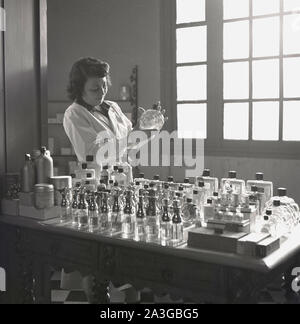 1950s, historical, Grasse, at a perfumery, a female chemist in a white-coat holding a bottle of Eau de Cologne, a bottle of perfurme, one of many different scents displayed on a table in the room. The French town is known as the perfume capital of the world. Stock Photo