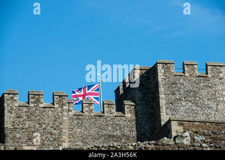 White bird about to land on Union Jack flag pole, flag flying on a sunny day over Dover Castle, Kent England. Rooftop crenelations of Castle Keep. Stock Photo