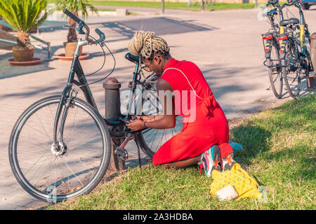 Valencia, Spain - September 28, 2019: Beautiful african girl Locking a bicycle to the post.  Girl in red dress channing her bike. View from behind. Stock Photo