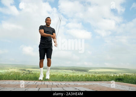 Handsome track and field athlete jumping outdoors. Man with sportive body in black t shirt and shorts training with jumping rope on background of nature and blue sky. Stock Photo