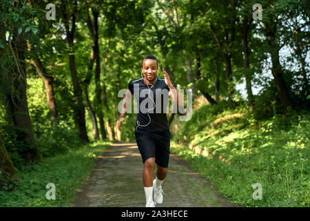 African athlete wearing in black t shirt and shorts running in park walkway. Handsome positive man listening music with headphones.