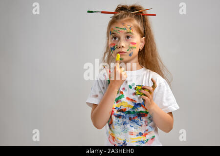 Little girl in white t-shirt, with brushes in her hair is posing standing isolated on white, gesticulating with painted hands and face. Close-up. Stock Photo