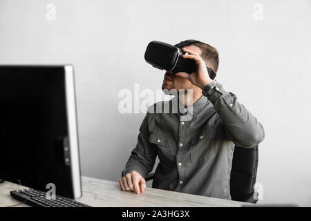 Young bearded man sitting against computer with virtual reality glasses. Modern technologies. The concept of future technology. Stock Photo