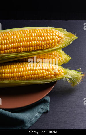 Three ripe corn ears with green leaves and plate on dark background Stock Photo