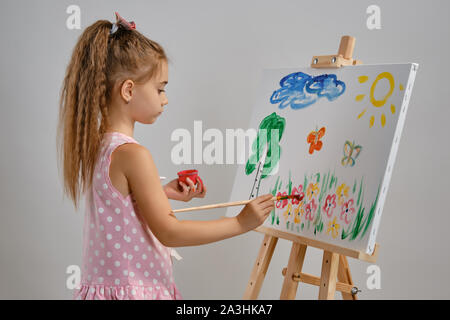 Little girl artist in a pink dress is standing behind easel and painting with brush on canvas, isolated on white studio background. Medium close-up. Stock Photo