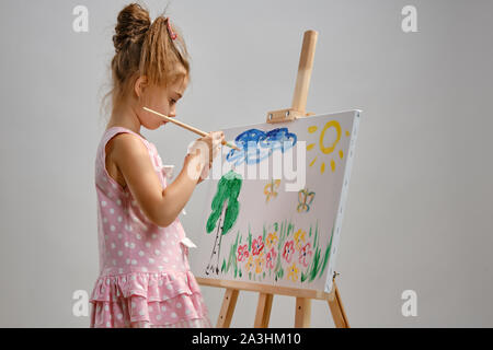 Little girl artist in a pink dress is standing behind easel and painting with brush on canvas, isolated on white studio background. Medium close-up. Stock Photo