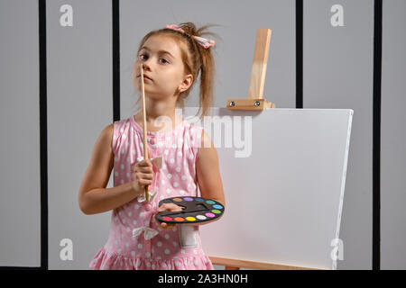 Little girl artist in a pink dress is standing behind easel and painting with brush on canvas at art studio with white walls. Medium close-up shot. Stock Photo