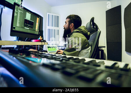 Young DJ with long beard is producing electronic music in his studio. Stock Photo