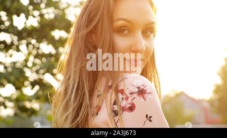 nice young woman with loose fair hair in pink dress smiles and poses for camera against bright sun close-up Stock Photo