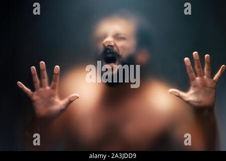 Creepy man looking and suffering behind the frosted glass and holding it with two hands. Stock Photo