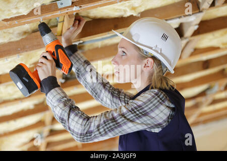 female builder using cordless drill on wooden ceiling joist Stock Photo