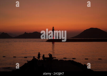 Low key panoramic image of a group of unrecognizable people in silhouettes, enjoying the dusk view of Aegean islands, calm sea, and a lighthouse. Stock Photo