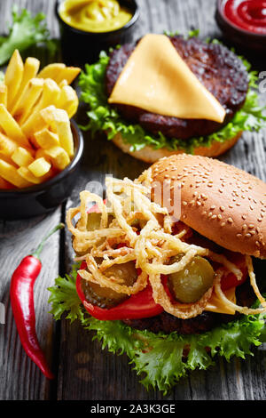 three Cheeseburgers with beef patties, cheddar cheese, crispy fried onions, lettuce, sliced tomatoes, pickles on a rustic wooden table with mustard an Stock Photo