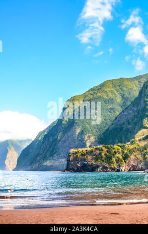 Amazing sand beach in Seixal, Madeira Island, Portugal. Green hills covered by tropical forests in the background. People swimming in the ocean. Vacation destination. Portuguese tourist attraction. Stock Photo