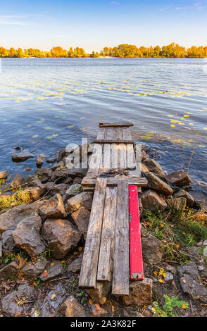 A wooden pontoon with a red paint plank  over big stones close to the blue Dnieper river is waiting for the fisherman. Stock Photo