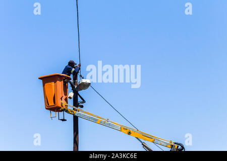 Electrician in crane bucket hydraulic arm extender high up  doing electrical light cable repairs maintenance on road  pole in blue sky. Stock Photo