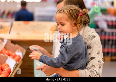 Cheerful smiling young woman with little daughter buying red tomatoes at the market. Mother and child choosing seasonal vegetables in supermarket. Fam Stock Photo