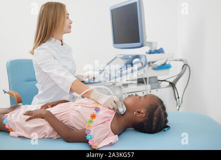 Pretty afro child lying on bed, during ultrasound of thyroid. Little girl, wearing in pink dress, courteously follows instructions of female doctor with ultrasound probe in hand and scanning her neck. Stock Photo