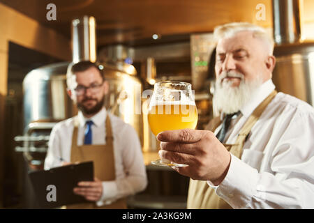 Bearded, elderly expert holding glass with light beer, looking at drink and testing. Concept of checking quality. Brewery workers examining drink in mini brewery. Stock Photo