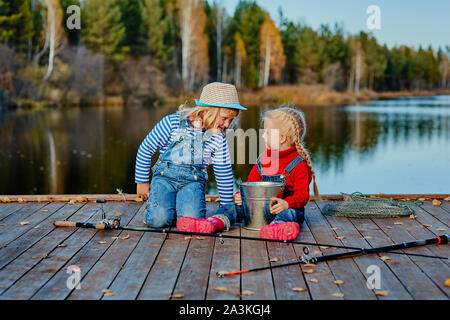 Two little sisters or friends sit with fishing rods on a wooden pier. They caught a fish and put it in a bucket. They are happy with their catch and d Stock Photo