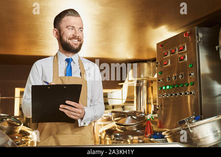 Handsome, cheerful brewery expert inspecting process of brewing beer. Engineer holding folder and pen. Positive worker in white shirt and brown apron smiling, looking away. Stock Photo
