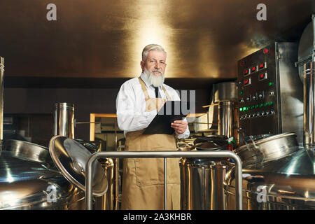 Professional brewery worker monitoring process of brewing beer. Handsome, elderly, bearded worker wearing in apron holding folder, looking at camera. Stock Photo