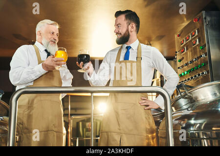 Two successful brewers standing on metal platform and holding beer glasses. Positive, cheerful brewery workers looking at each other, posing and smiling. Stock Photo