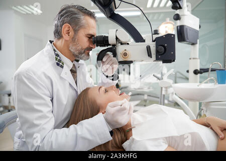 Side view of stomatologist using professional equipment for teeth diagnosis. Mature man working as dentist in private dentistry clinic. Patient lying on dentist chair. Stock Photo