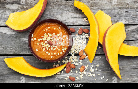 Pumpkin porridge or cream soup in a bowl decorated with oat flakes and almond, slices of cut squash are nearby on wooden rustic background from above, Stock Photo