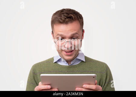 Handsome surprised young man wearing green sweater looking on tablet Stock Photo