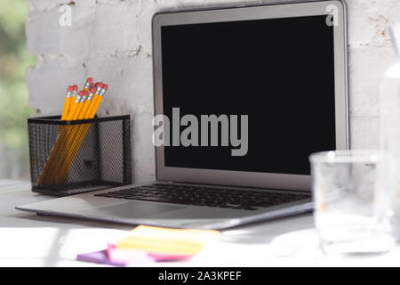 Mock up empty black laptop screen on white brick wall background with pencils, notes and glass of water. Copyspace, negative space for your advertisin Stock Photo