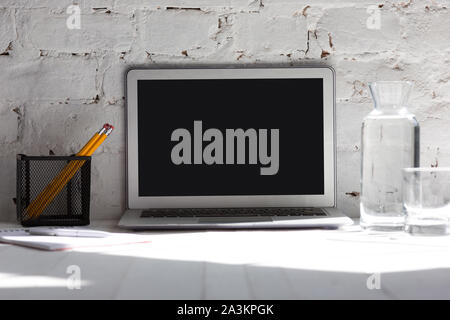 Mock up empty black laptop screen on white brick wall background with pencils, notes and glass of water. Copyspace, negative space for your advertisin Stock Photo