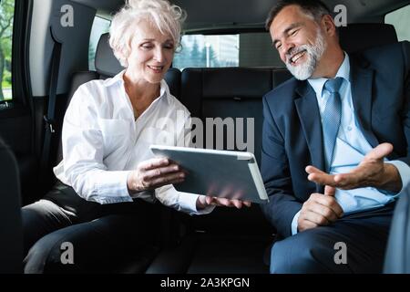Senior business colleagues working on document in back seat of car. Businessman and businesswoman using tablet and preparing for meeting. Stock Photo