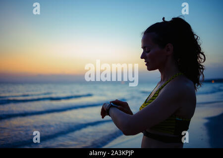 Silhouette of healthy sports woman in sport style clothes on the seacoast in the evening using smart watch to track heart rate in fitness app. Stock Photo