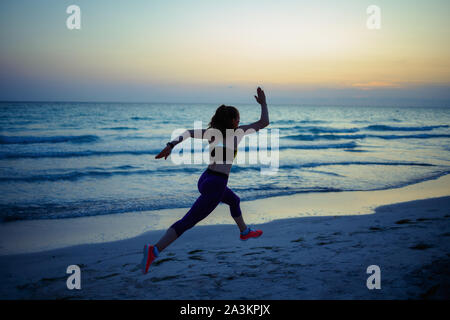 Seen from behind fit sports woman in sport style clothes on the ocean shore in the evening jogging. Stock Photo