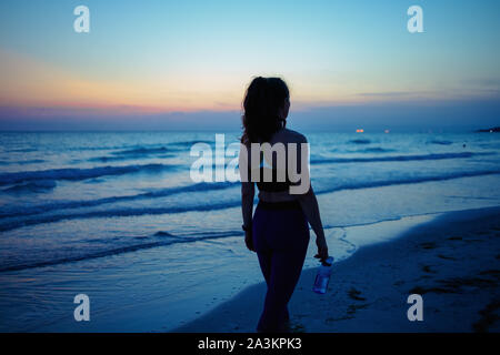 Seen from behind young woman in fitness clothes with bottle of water walking after workout on the ocean coast in the evening. Stock Photo