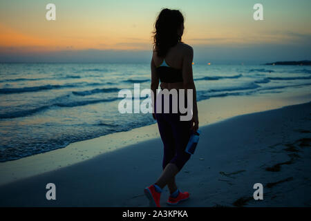 Seen from behind fitness sports woman in fitness clothes with bottle of water walking after workout on the ocean shore in the evening. Stock Photo