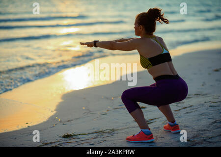 Seen from behind young sports woman in sport clothes on the beach in the evening doing squats. Stock Photo