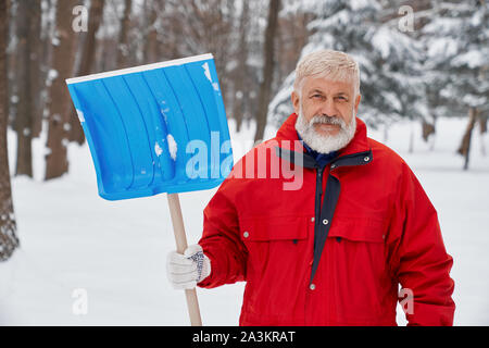 Front view of elder man wearing uniform keeping shovel and looking at camera while cleaning snow from streets. Male janitor posing while working in morning in winter. Concept of maintenance. Stock Photo