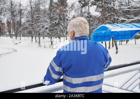 View from back of grey bearded man in blue uniform leaning on handrails, keeping shovel on shoulder and looking forward in winter. Male resting after cleaning footpath from snow. Concept of service. Stock Photo